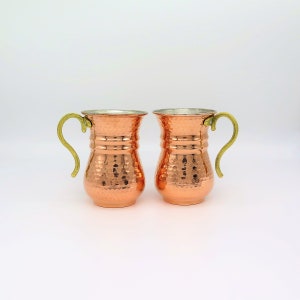 May include: Two copper mugs with a hammered texture and brass handles. The mugs have a flared rim and a shiny finish. The handles are curved. The mugs are displayed on a white background. Each mug is approximately 10 cm tall.