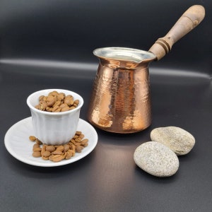 May include: A copper coffee pot with a wooden handle and a white ceramic cup filled with coffee beans. The coffee pot has a hammered texture and the cup is sitting on a white saucer with more coffee beans.