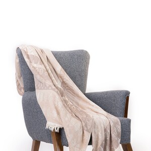 Puede incluir: Un sill&oacute;n gris con patas de madera tiene una manta beige drapeada sobre &eacute;l. La manta tiene un patr&oacute;n sutil y un ribete de flecos.