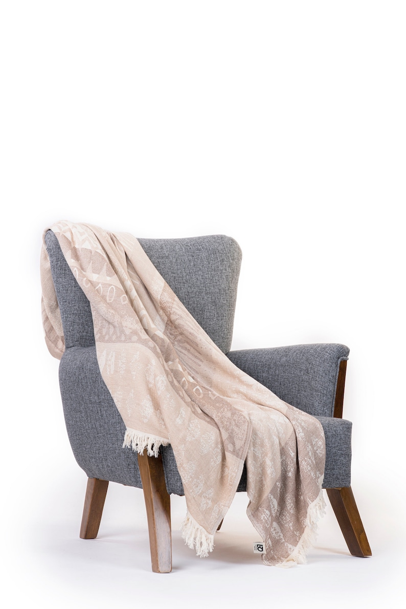 Puede incluir: Un sill&oacute;n gris con patas de madera tiene una manta beige drapeada sobre &eacute;l. La manta tiene un patr&oacute;n sutil y un ribete de flecos.
