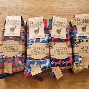 May include: Four pairs of patterned socks featuring alpaca and merino wool blends. Each pair has a brown cardboard label with the text Mit ALPAKA & MERINO Fuß-Schmeichler and an alpaca illustration. The socks display colourful geometric and animal motifs.