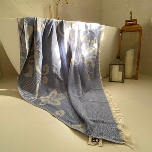 Puede incluir: Una toalla turca de color azul claro con estampado y bordes con flecos, drapeada sobre el borde de una ba&ntilde;era blanca. La toalla tiene un logotipo blanco que dice "LEO DESIGN Istanbul".