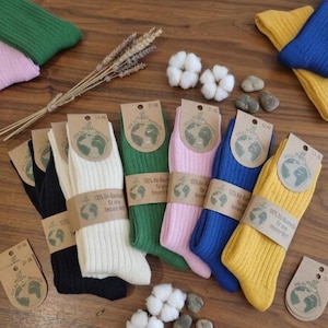 May include: A collection of ribbed socks in black, white, green, pink, blue, and yellow, each secured with a brown paper label featuring a globe graphic and the text 100% Bio-Baumwolle für eine bessere Welt. Cotton bolls and stones are scattered nearby.