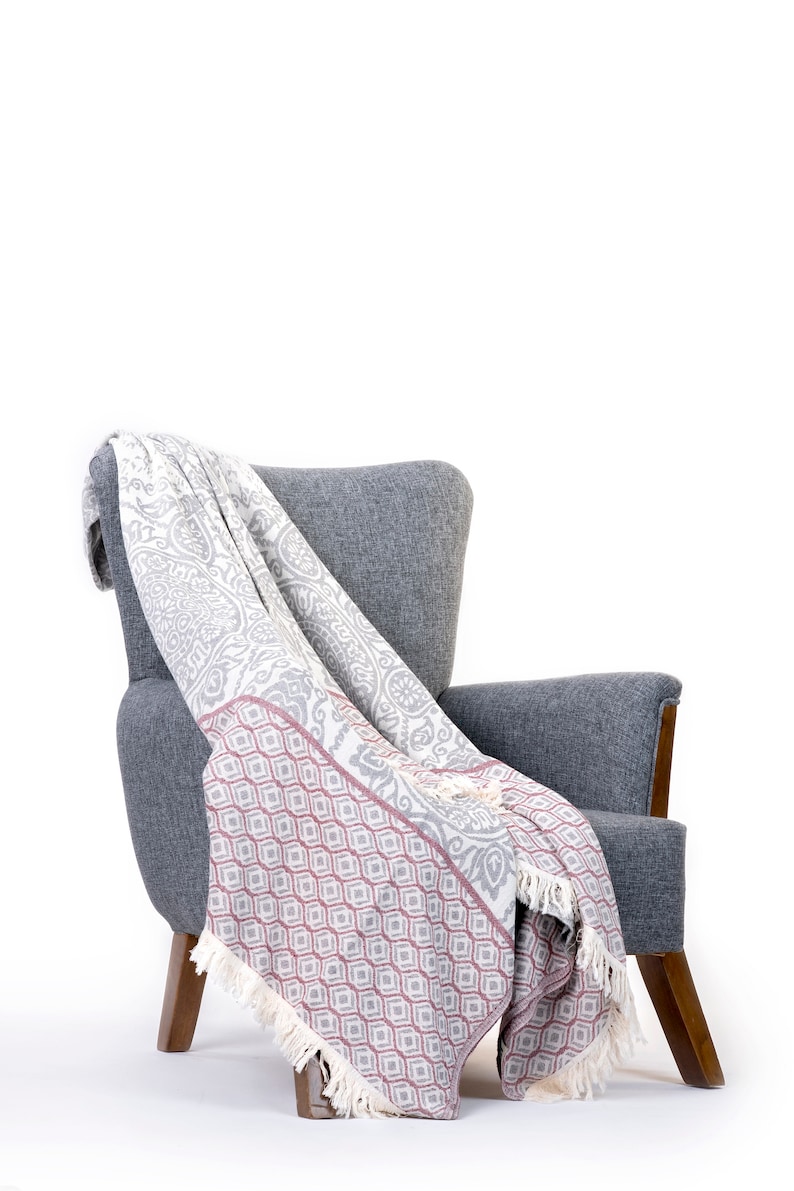 Puede incluir: Un sill&oacute;n gris con una manta de tiro blanca y gris drapeada sobre &eacute;l. La manta tiene un patr&oacute;n geom&eacute;trico rosa y blanco en la mitad inferior.