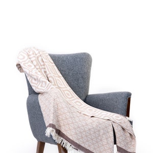 Puede incluir: Un sill&oacute;n gris con una manta de tiro beige y blanca con estampado drapeada sobre &eacute;l. La manta tiene un borde de flecos.