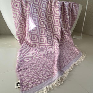 Puede incluir: Una toalla turca con flecos de color morado y blanco. La toalla tiene un dise&ntilde;o geom&eacute;trico con fondo blanco y detalles morados.