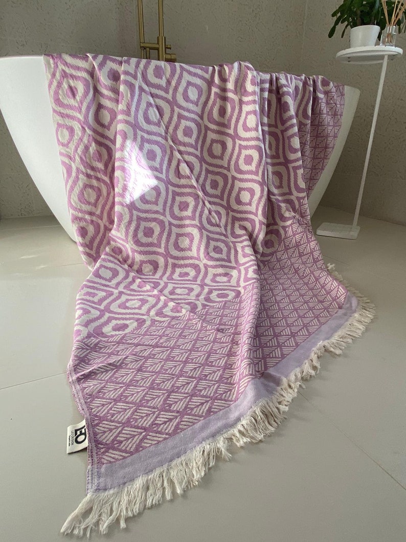 Puede incluir: Una toalla turca con flecos de color morado y blanco. La toalla tiene un dise&ntilde;o geom&eacute;trico con fondo blanco y detalles morados.