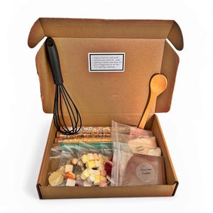 May include: A cardboard box filled with baking ingredients. A black whisk and wooden spoon are included. Inside are packets of ingredients, such as chocolate powder, sprinkles, and marshmallows. A small white label with text is affixed to the box.