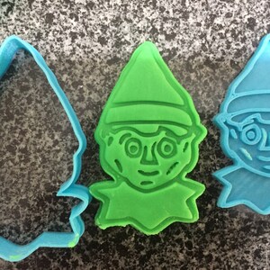 May include: Two blue and one green plastic cookie cutters shaped like a gnome with a pointed hat. The gnome has large eyes, a nose, and a mouth.