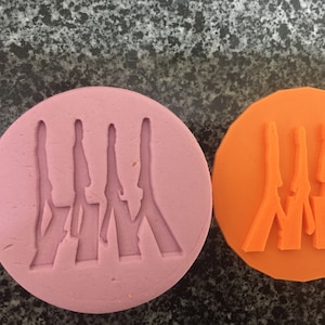 May include: Two round silicone moulds for baking, one pink and one orange. Both moulds feature a design of four figures walking across a street, similar to the iconic Abbey Road album cover.
