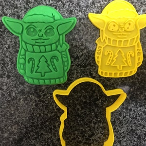 Può includere: Due stampini per biscotti in plastica verde e gialla a forma di un personaggio che indossa un maglione natalizio. Il maglione ha un albero di Natale e bastoncini di zucchero.