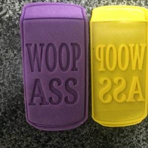 Puede incluir: Dos cortadores de galletas con forma de latas de aluminio. Uno es morado y dice "Woop Ass" y el otro es amarillo y dice "Woop Ass".
