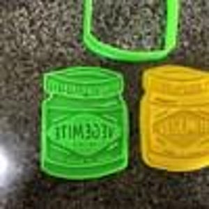 May include: Green and yellow plastic cookie cutters shaped like jars with lids. The green cutter has the word "VEGEMITE" printed on it. The cutters are on a speckled surface.