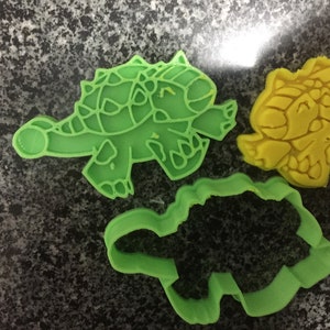 Puede incluir: Dos cortadores de galletas de dinosaurio verde y un cortador de galletas de dinosaurio amarillo. Los cortadores verdes están detallados con escamas y garras. El cortador amarillo es más estilizado.