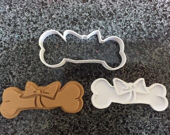May include: Two white plastic dog bone cookie cutters and a brown dog bone cookie. The cookie cutters have a bow on top.