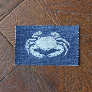 May include: A rectangular denim patch featuring a white crab design. The crab is centered on the blue fabric, with its claws and body clearly visible. The patch is placed on a wooden surface.