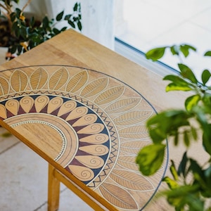 Może przedstawiać: Drewniany stolik z ręcznie malowanym wzorem mandali w kolorze złotym, brązowym i czarnym. Wzór przedstawia półkole z liści i spiral.