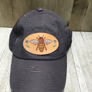 May include: A grey distressed baseball cap with a brown leather patch featuring a yellow and brown bee with blue flowers.