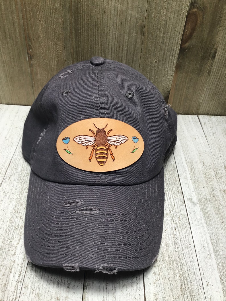 May include: A grey distressed baseball cap with a brown leather patch featuring a yellow and brown bee with blue flowers.