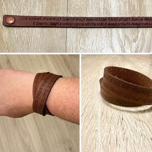 Op de afbeelding: Bruine leren armband met gegraveerde tekst. De armband wordt plat en om een pols getoond. De tekst luidt "Liefde is geduldig, liefde is vriendelijk..." en andere zinnen. Een knoopsluiting is zichtbaar.