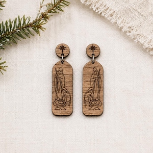 May include: A pair of wooden earrings featuring a Nativity scene. Each earring has a circular top with a star design and a rectangular pendant with an engraved depiction of Mary, Joseph, and baby Jesus. The earrings are a light brown color.