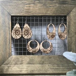 Peut inclure: Quatre paires de boucles d'oreilles en bois avec des motifs de tournesols. Les boucles d'oreilles sont expos&eacute;es sur un rack en fil de fer sur un fond en bois.