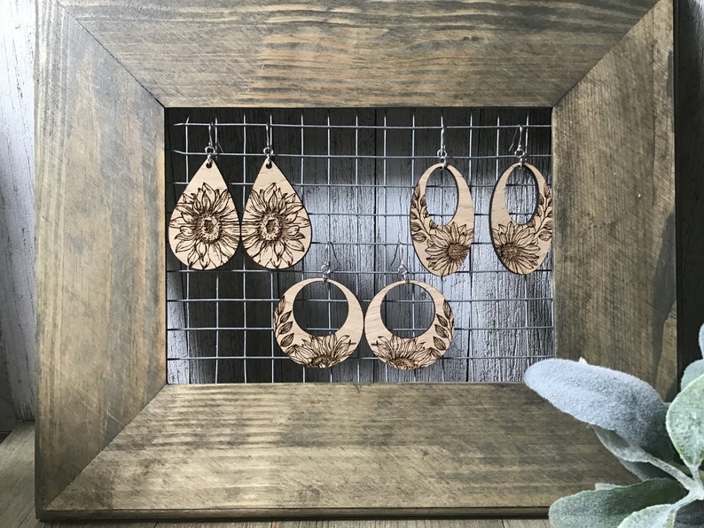 Peut inclure: Quatre paires de boucles d'oreilles en bois avec des motifs de tournesols. Les boucles d'oreilles sont expos&eacute;es sur un rack en fil de fer sur un fond en bois.