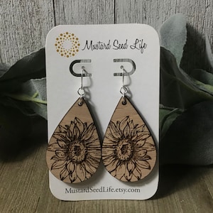 Peut inclure: Une paire de boucles d'oreilles en bois en forme de goutte d'eau avec un motif de tournesol grav&eacute; au laser. Les boucles d'oreilles sont sur une carte blanche avec le texte "Mustard Seed Life" et "MustardSeedLife.etsy.com".