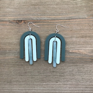 May include: A pair of teal and light blue acrylic earrings with a geometric arch shape. The earrings have a silver hook.