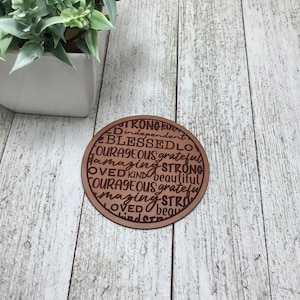May include: A brown leather coaster with the words "STRONG, BLESSED, COURAGEOUS, LOVED, KIND, AMAZING, GRATEFUL, INDEPENDENT, BEAUTIFUL" engraved in a circular pattern.