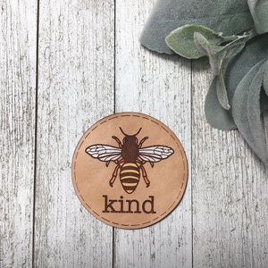 Peut inclure: Un dessous de verre rond en cuir brun avec un bord cousu présente une abeille de dessin animé avec des rayures jaunes et noires et le mot "kind" imprimé en dessous.