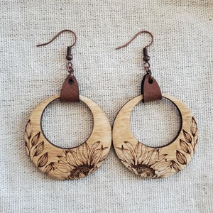 Peut inclure: Une paire de boucles d'oreilles en bois en forme de cercle avec un motif de tournesol. Les boucles d'oreilles sont en bois naturel et ont un accent en cuir brun.