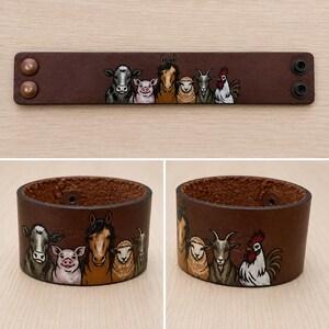 Pode incluir: Uma pulseira de couro castanho com um desenho pintado de animais de fazenda. Os animais incluem uma vaca, um porco, um cavalo, uma ovelha, uma cabra e um galo. A pulseira tem fechos de pressão.
