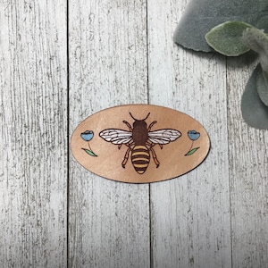 Peut inclure: Un patch en cuir ovale de couleur marron avec une abeille et deux fleurs bleues. L'abeille est jaune et noire avec des ailes brunes. Les fleurs ont des tiges vertes et des pétales bleus.