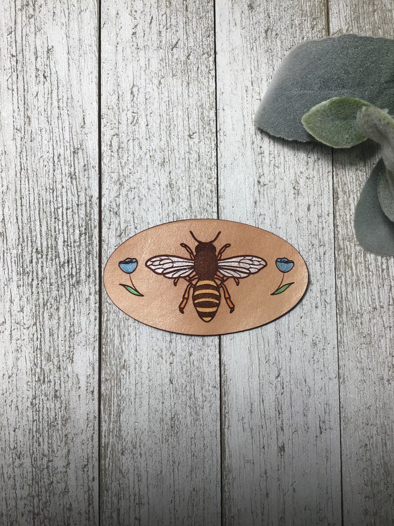 May include: An oval-shaped leather patch in brown, featuring a bee and two blue flowers. The bee is yellow and black with brown wings. The flowers have green stems and blue petals.