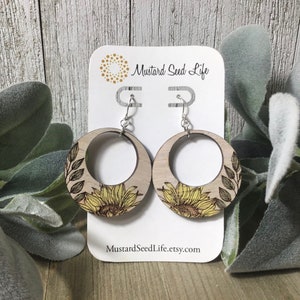 Peut inclure: Une paire de boucles d'oreilles en bois en forme de cercle avec un motif de tournesol. Les boucles d'oreilles sont de couleur brun clair avec un tournesol jaune et des feuilles vertes. Les boucles d'oreilles sont sur une carte blanche avec le texte "Mustard Seed Life" et "MustardSeedLife.etsy.com".