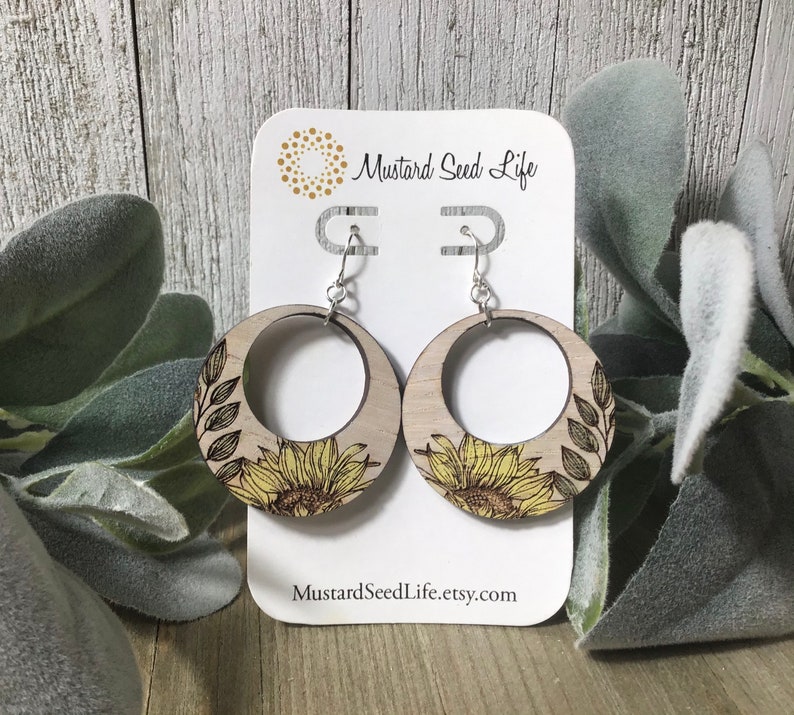 Peut inclure: Une paire de boucles d'oreilles en bois en forme de cercle avec un motif de tournesol. Les boucles d'oreilles sont de couleur brun clair avec un tournesol jaune et des feuilles vertes. Les boucles d'oreilles sont sur une carte blanche avec le texte "Mustard Seed Life" et "MustardSeedLife.etsy.com".