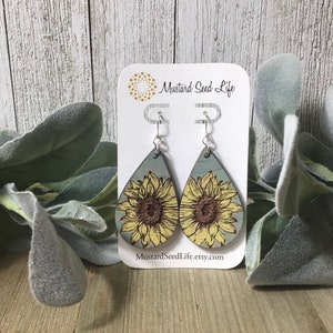 Peut inclure: Une paire de boucles d'oreilles en bois en forme de goutte d'eau avec un motif de tournesol. Les boucles d'oreilles sont peintes dans des tons de bleu, jaune et marron. Les boucles d'oreilles sont suspendues &agrave; des crochets argent&eacute;s. Les boucles d'oreilles sont pr&eacute;sent&eacute;es sur une carte blanche avec le texte "Mustard Seed Life" et "MustardSeedLife.etsy.com".