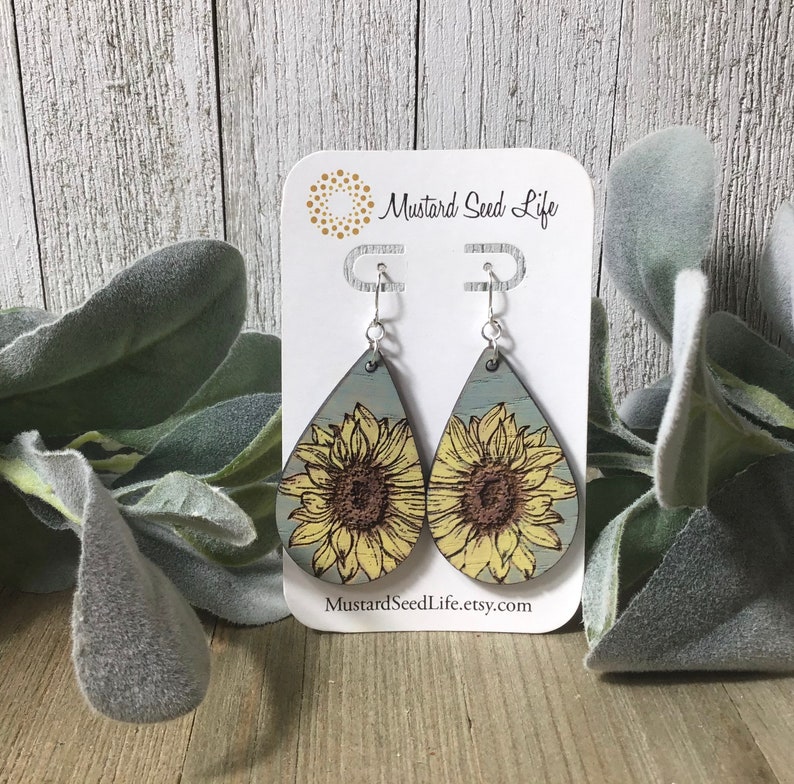 Peut inclure: Une paire de boucles d'oreilles en bois en forme de goutte d'eau avec un motif de tournesol. Les boucles d'oreilles sont peintes dans des tons de bleu, jaune et marron. Les boucles d'oreilles sont suspendues &agrave; des crochets argent&eacute;s. Les boucles d'oreilles sont pr&eacute;sent&eacute;es sur une carte blanche avec le texte "Mustard Seed Life" et "MustardSeedLife.etsy.com".