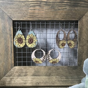 Peut inclure: Quatre paires de boucles d'oreilles en bois avec des motifs de tournesols. Les boucles d'oreilles sont expos&eacute;es sur une grille m&eacute;tallique sur un fond en bois rustique.