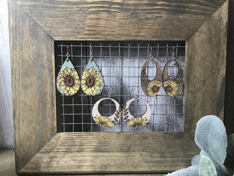 Peut inclure: Quatre paires de boucles d'oreilles en bois avec des motifs de tournesols. Les boucles d'oreilles sont expos&eacute;es sur une grille m&eacute;tallique sur un fond en bois rustique.