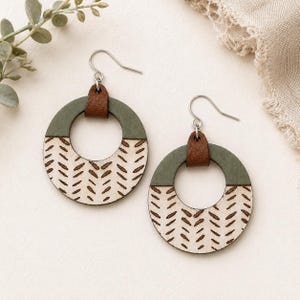 May include: A pair of wooden hoop earrings. The top section is olive green, connected to a brown leather strap and silver hook. The bottom half is a natural wood color with a brown, patterned design. The earrings are displayed on a white surface.