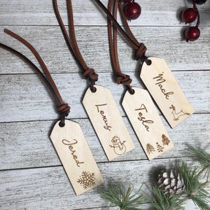 May include: Four wooden gift tags with brown leather cords. The tags are engraved with the names "Mack", "Tesla", "Lexy", and "Jered" and feature a snowflake, snowman, Christmas tree, and reindeer design.
