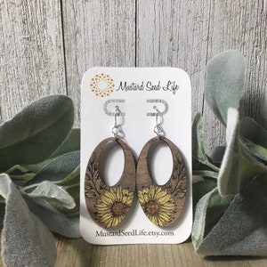 Peut inclure: Une paire de boucles d'oreilles en bois avec un motif de tournesol. Les boucles d'oreilles sont de forme ovale et ont un crochet argent&eacute;. Les boucles d'oreilles sont sur une carte blanche avec le texte "Mustard Seed Life" et "MustardSeedLife.etsy.com".