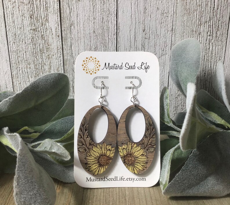Peut inclure: Une paire de boucles d'oreilles en bois avec un motif de tournesol. Les boucles d'oreilles sont de forme ovale et ont un crochet argent&eacute;. Les boucles d'oreilles sont sur une carte blanche avec le texte "Mustard Seed Life" et "MustardSeedLife.etsy.com".