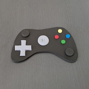 May include: A black games controller with a grey cross-shaped directional pad, a white button, and coloured buttons. The buttons are yellow, red, green, and blue. The controller is on a grey surface.