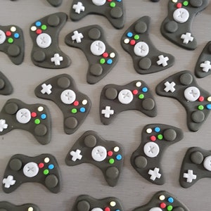 May include: A collection of black and grey fondant video game controllers with colourful buttons. The controllers are arranged in a grid pattern on a grey surface.