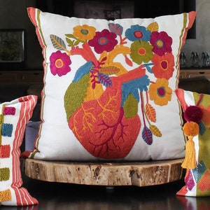 May include: Four colourful embroidered throw pillows with various patterns, including stripes, hearts, and a floral design with a heart. The pillows are arranged on a wooden surface.