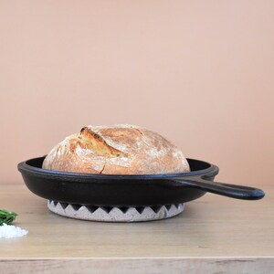 May include: A loaf of freshly baked bread sits in a black cast iron skillet on a gray and white trivet. The skillet has a long handle.