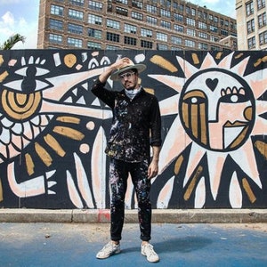 May include: A person wearing a black shirt and a hat stands in front of a large mural. The mural features a black, white, and gold design with a sun and a bird-like creature.